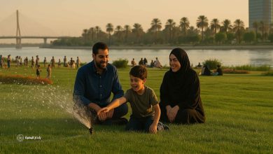 أم بصرية شابة وأب بصري شاب يجلسان على العشب الأخضر مع طفلهما الذي يلعب بمرح مع قطرات الماء في حديقة على كورنيش البصرة وقت الغروب، في مشهد يعكس الأمل والرخاء.